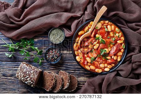 Hearty Beans Stew With Sausages, Herbs And Spices In Tomato Sauce In A Black Bowl With Spoon, On A R