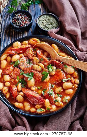 Beans Stew With Sausages, Herbs And Spices In Tomato Sauce In A Black Bowl With Spoon, On A Rustic W