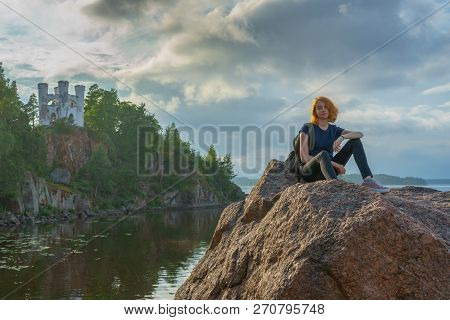Pretty Young Lady With Red Hair Sitting On Stone Near Bay Shore In Summer Evening. Tourist On The Be