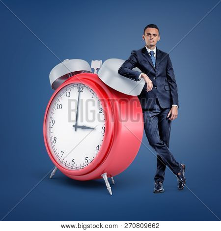 A Small Businessman Stands Looking At Front And Leaning On A Huge Red Alarm Clock.