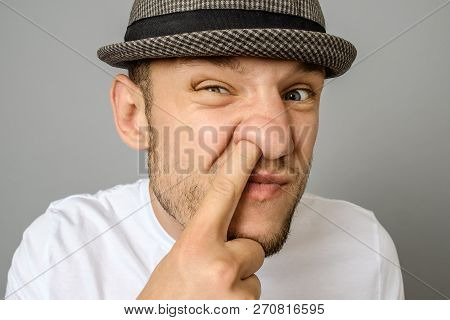 Man Picking His Nose. The Emotional Portrait Of Man In Hat And Blank T-shirt. Gray Background
