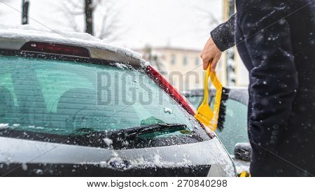 Young Man In Black Coat Cleans His Car With Yellow Brush During Snowfall. Winter Inclement Weather.
