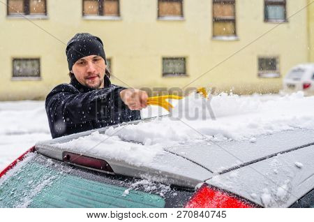 Young Man In Black Coat Cleans His Car With Yellow Brush During Snowfall. Winter Inclement Weather.