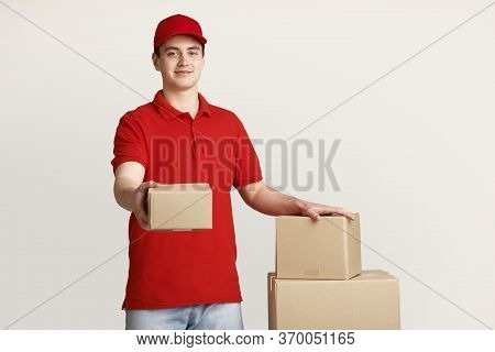 Issuance Of Parcels At Post Office. Courier With Red Uniform Holds Out Box And Puts His Hand On Stac