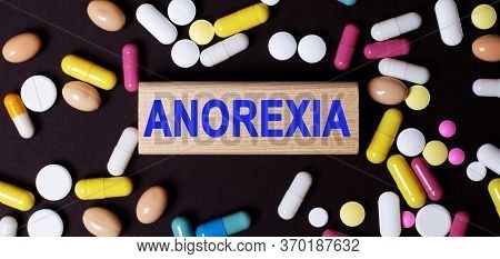 Wooden Block With The Word Anorexi On A Dark Background Among Multi-colored Pills. Anorexia Nervous 