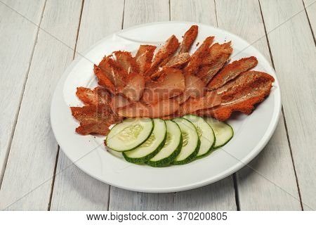 Meat smoked jerky - raw meat . Close up view on tasty sliced Chicken basturma with slices of cucumber on white plate on light wooden background. Beer snack. Pastirma