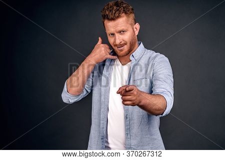 Young Attractive Guy With Red Hair In Casual Shirt, Makes Phone Sign, Indicates With Index Finger Di