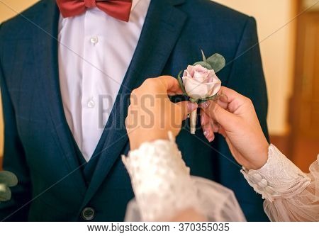 Beautiful Bride Puts A Flower On The Groom's Jacket In A Hotel Room. White Dress And Blue Suit.