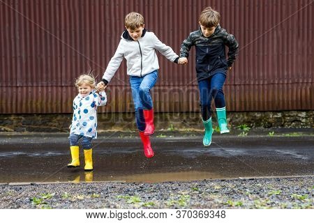 Three Children, Toddler Girl And Two Kids Boys Wearing Red, Yellow And Green Rain Boots And Walking 