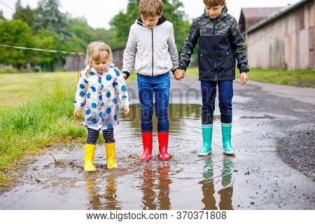 Three Children, Toddler Girl And Two Kids Boys Wearing Red, Yellow And Green Rain Boots And Walking 