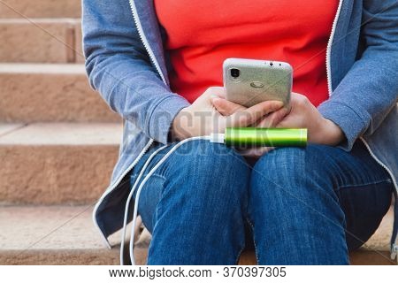 A Girl Sitting On The Steps Uses A Smartphone Outdoors While Charging From An External Power Bank