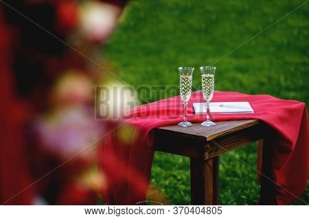 Champagne Glasses On Red Table At Evening Wedding Ceremony Reception, With Lights On Background. Spa