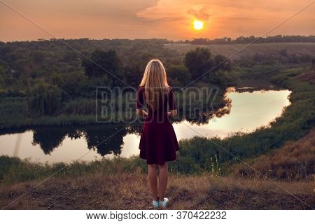 Young Blond Girl In Beautiful Burgundy Dress And Hat Have Fun At Park Near Water