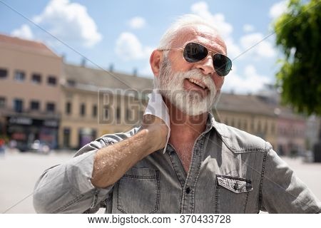 Old man cleaning neck and sweat with wet wipes on street on hot summer day, blurred urban city background