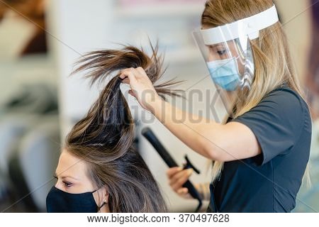 Hairdresser, Protected By A Mask, Combing Her Clients Hair With A Hair Iron In A Salon.
