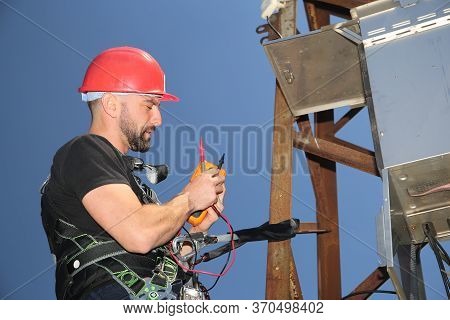Tehnician In Red Helmet And Safety Belt With Multimeter On The Electric Pole