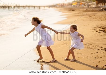 Girl Children In White Dresses Playing On The Beach. Older Sister Pulls The Younger Into The Water. 