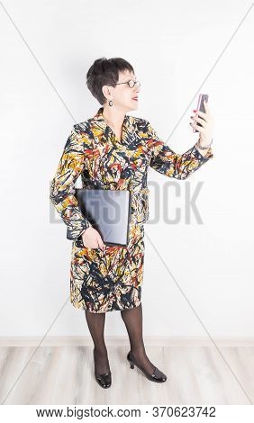 Senior Woman Holding Laptop Computer Isolated On Grey Studio Background. She Looking At Phone