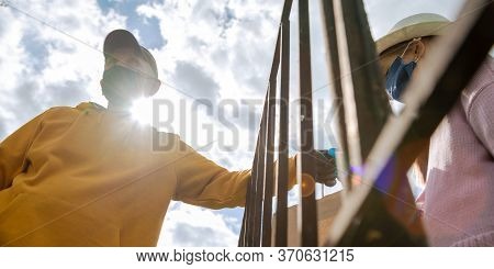 Pensioner And Girl Hands In Gloves Hold Paper Bag With Products Over Metal Fence Against Sky At Back