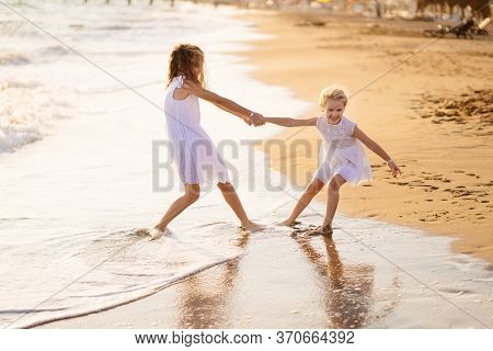 Girl Children In White Dresses Playing On The Beach. Older Sister Pulls The Younger Into The Water. 