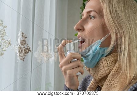 Blonde Girl With A Lowered Mask On Her Chin Is Spraying Throat Spray While Standing Near The Window.