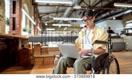 Portrait Of Young Male Office Worker In A Wheelchair Looking At The Screen Of His Laptop While Perfo