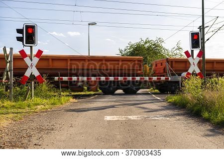 Closed Barrier At The Railroad Crossing With Red Warning Lights On, Visible Blurred Red Wagons In Mo