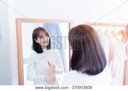 Asian Woman Choosing Make Up With Cosmatic Applying Lipstick And Smiling Happy In Front Mirror At Ho