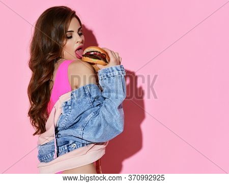 Sexy Curly Brunette Woman In Pink Bodysuit And Denim Jacket Eats Fresh Fast Food Hamburger Burger Ov