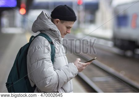 Side View Of Young Man In Casual Gray Jacket And Hat Using Smartphone While Waiting Train At Railway