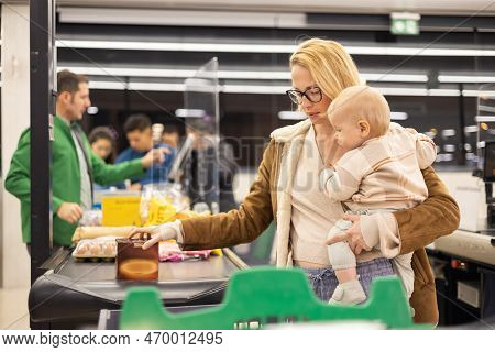 Mother Shopping With Her Infant Baby Boy, Holding The Child While Stacking Products At The Cash Regi