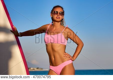 Tanned Girl Model Of Athletic Build In A Pink Swimsuit Poses On The Beach Against The Blue Sky On A 
