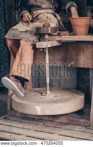 Craftsman In Dirty Working Clothes Sit On Potter Machine And Spin Wheel With Leg. Make Crockery In P