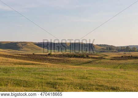 A Grassy Field With Hill With Forests In The Distance. Rural Scene, Country Road And Old Farm In Cou
