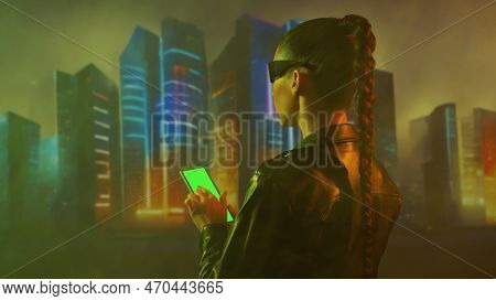 Portrait Of Cyberpunk Girl With A Smartphone. Beautiful Young Woman On The Background Of City Scyscr