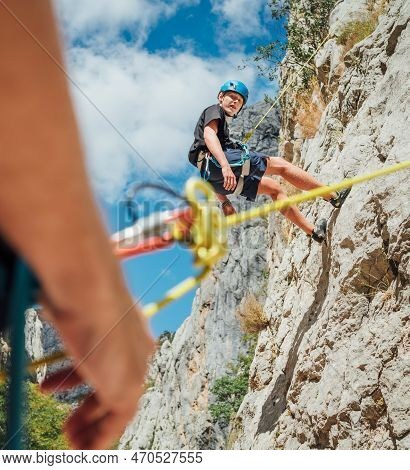Teenager Boy On The Natural Cliff Climbing Wall. Boy Hanging On A Rope Using A Climbing Harness And 