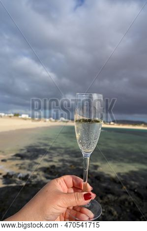 Hand With Glass Of White Champagne Or Cava Sparkling Wine On White Sandy Tropical Beach And Blue Oce