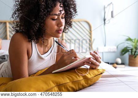 Pensive Young Multiracial Latina Woman Lying Down On Bed With Serious Expression Writing On Journal 