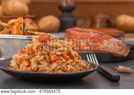 Stewed Cabbage - Bigos In A Black Plate On The Background Of A Piece Of Smoked Meat And Baked Potato
