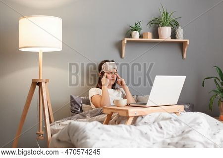 Horizontal Shot Of Tired Sick Unhealthy Woman Wearing Sleeping Mask Posing In Front Of Laptop At Hom