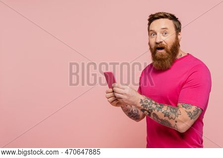 Excited Bearded Man With Open Mouth Using Cellphone And Looking At Camera Isolated On Pink.