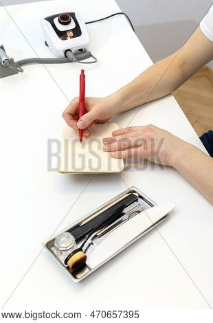 Unrecognizable Nail Master Makes Notes In Notebook With Pen, Sitting At Workstation, Table In Nail B