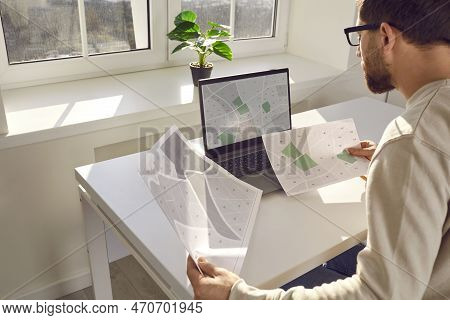Serious Engineer With Cadastral Maps And Projects Is Sitting At Desk With Laptop In Office. Cadastra