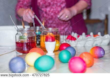 Old Woman Dyeing Easter Eggs At Home In The Kitchen In The Cucumber Glass And With Different Colors,