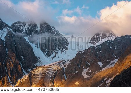Scenic Mountain Landscape With Snowy Mountain Top And Glacier In Low Clouds In Golden Sunrise Colors