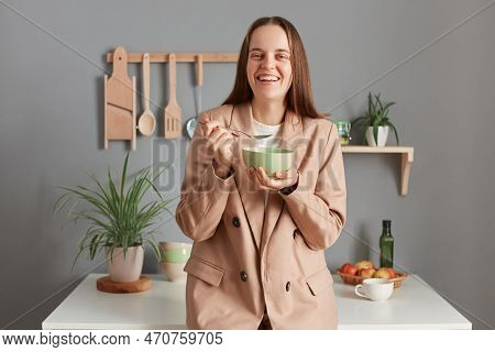 Indoor Shot Of Smiling Delighted Brown Haired Woman Wearing Beige Suit Standing Near Table On Kitche