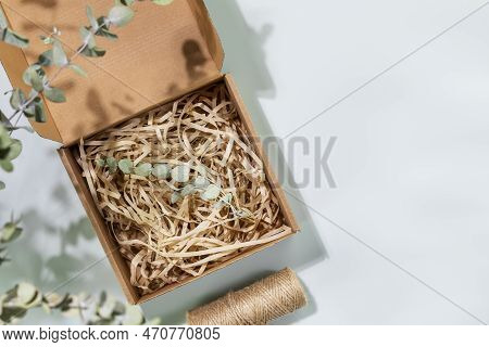 Eco Friendly Packaging Concept. Cardboard Box With Shredded Paper And Eucalyptus Sprig On Blue Backg