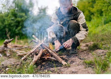 Close-up Of Blurred Survivalist Male In Raincoat Starting Fire Using Gas Lighter On Bank Of River Fo