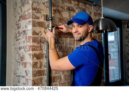 Young Plumber Dressed In A Blue Uniform And A Blue Cap Is Working With A Wrench To Repair A Pipe In 