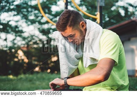 Young Man Looking Down At His Sports Watch After His Training, While Wiping Away The Sweat